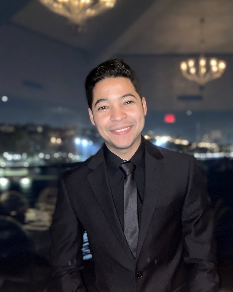 Portrait of Wilfre Garcia, founder of FinanzaX: a smiling man in a black suit and tie, photographed from above the head to mid-torso against a softly lit window.